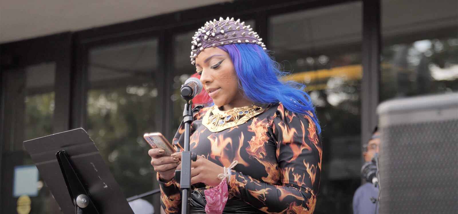 Video still of artist Abigail DeVille, with blue and purple hair, a large gold necklace, and a dress covered in flames, standing in front of a microphone in the Hirshhorn Museum’s sculpture garden