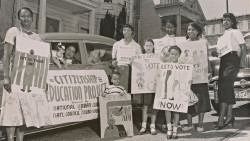 A black-and-white photograph of young children and members of the San Francisco Chapter of the National Council of Negro Women with placards standing next to a car that was part of the Citizenship Education Project motorcade urging people to register to vote